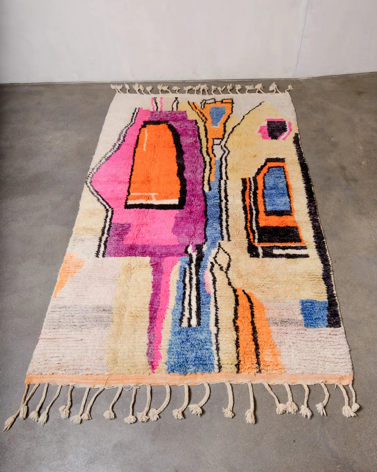 Handwoven Berberlin Berber rug from Morocco laying flat on concrete studio floor. Vintage carpet featuring a modern design in pink, yellow, orange, blue and black wool.